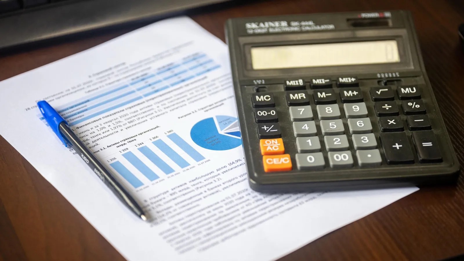 A desktop calculator resting on a printed financial report with bar and pie charts, next to a pen, on a wooden desk.