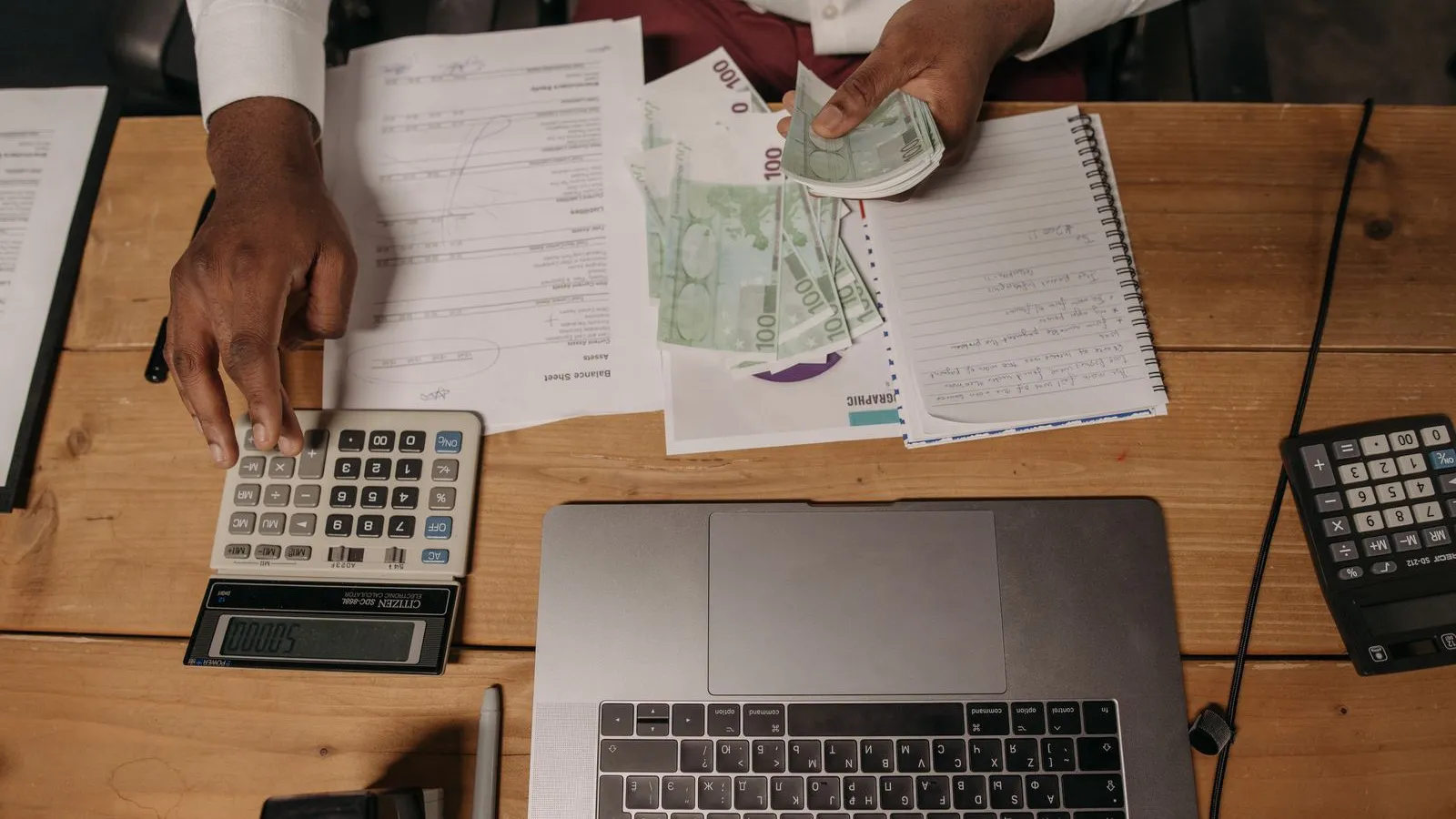 Overhead view of a desk with a calculator, banknotes, financial documents and a laptop, someone counting cash and using the calculator.