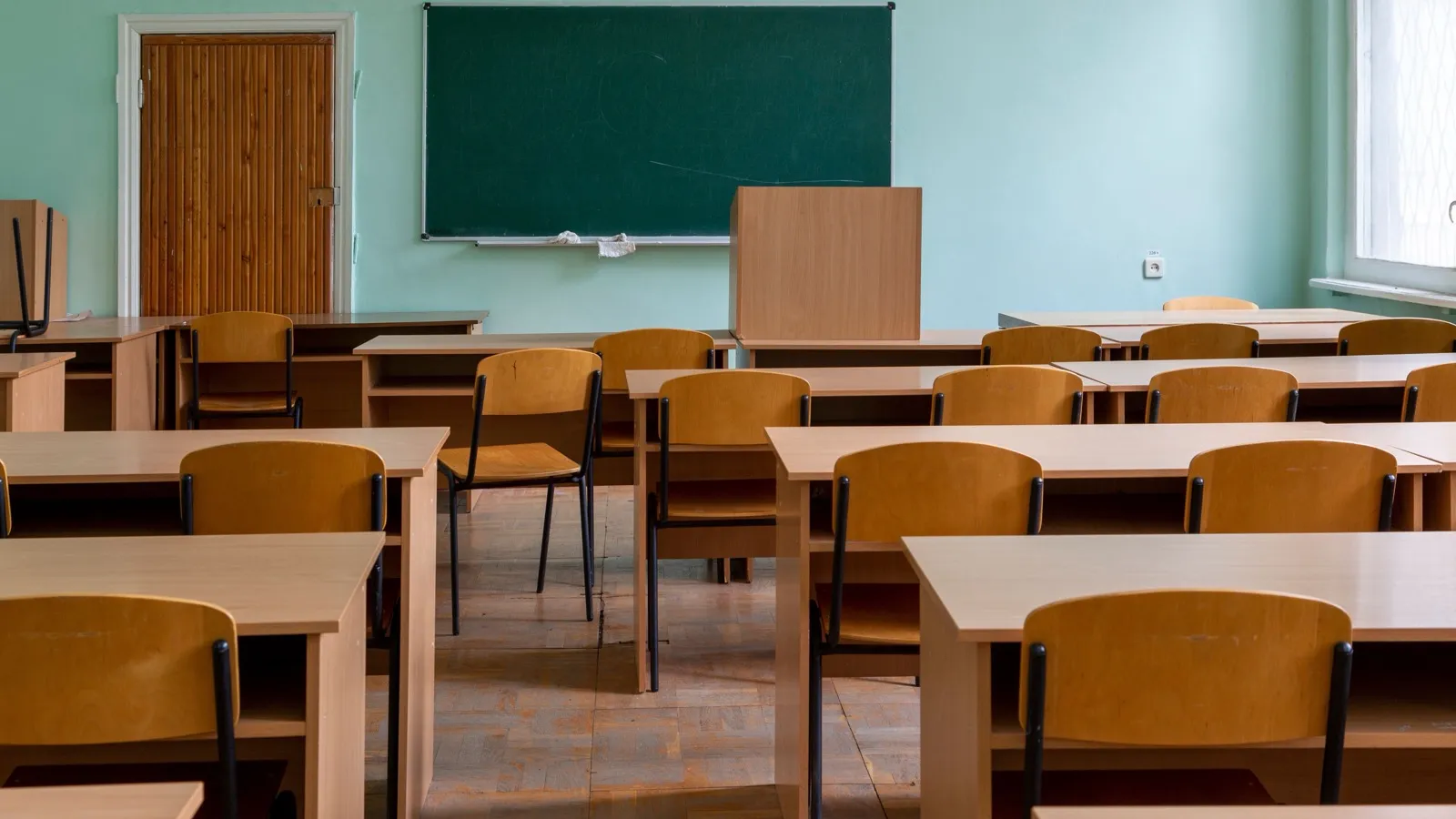 An empty classroom with rows of vacant wooden desks and chairs, a blank green chalkboard and a closed door, no students present.