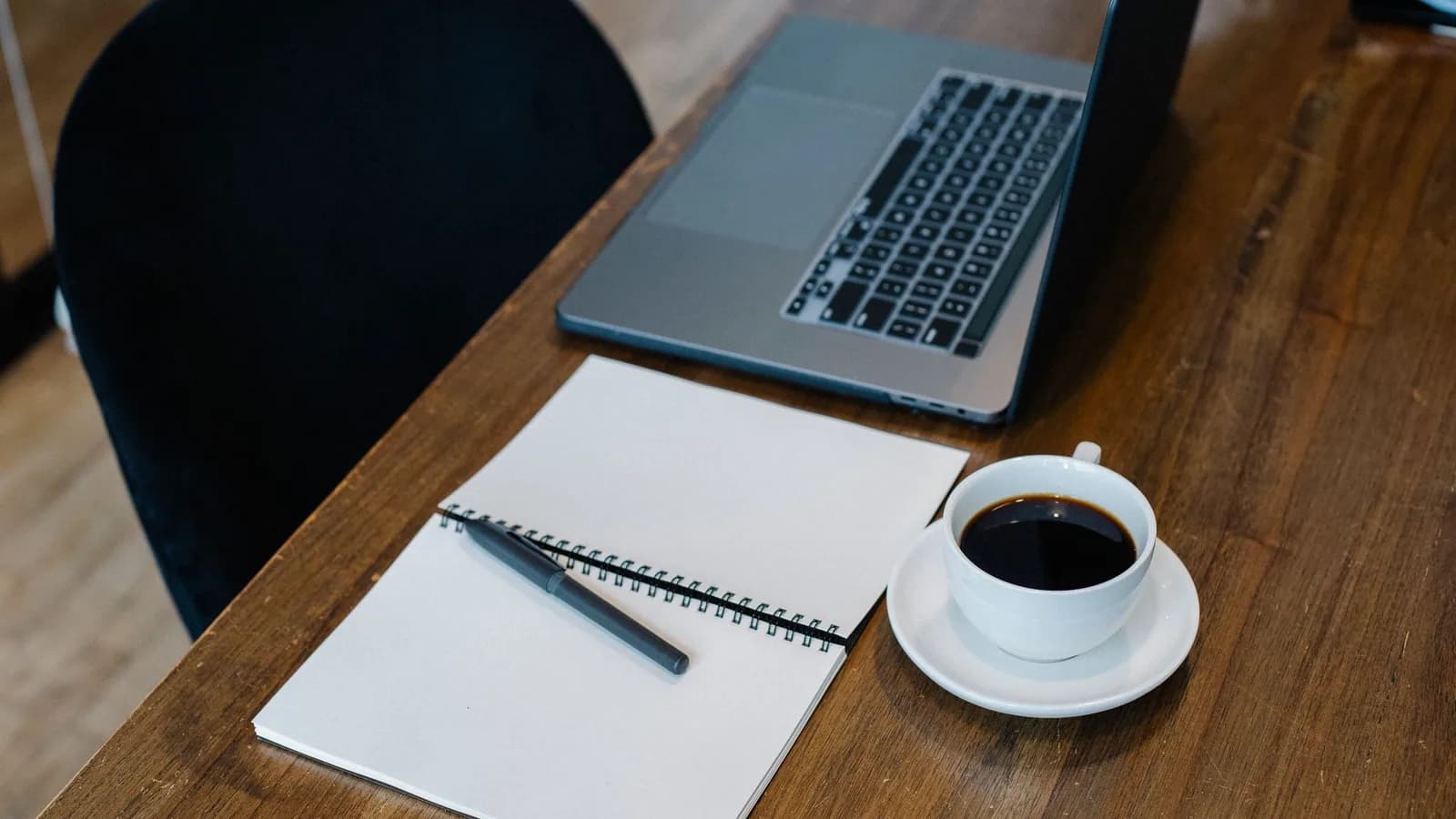 A laptop, open notebook and coffee on a wooden desk.