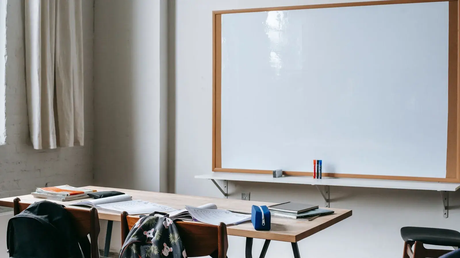 School bags, books and stationery on desks beneath a whiteboard.
