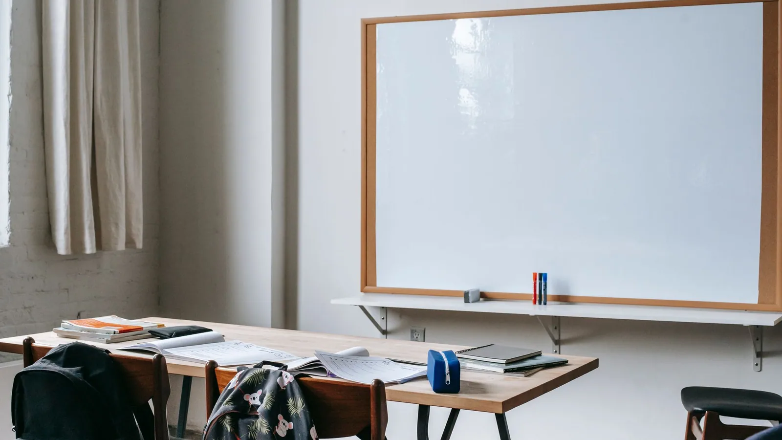 School bags, books and stationery on desks beneath a whiteboard.