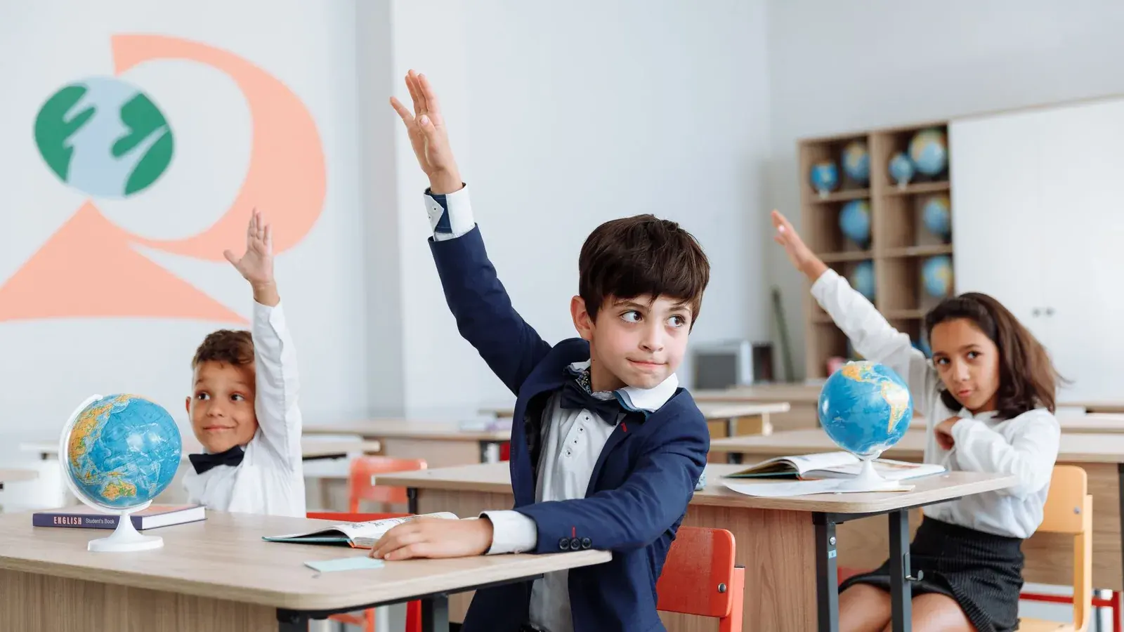 Children raising their hands at classroom desks with books and globes.