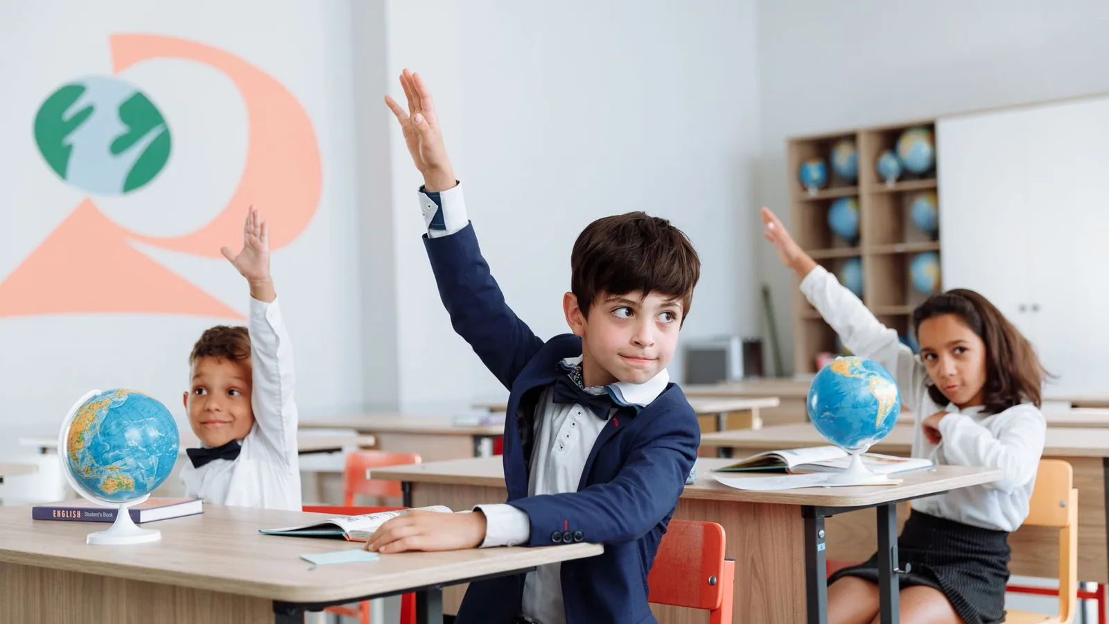 Children raising their hands at classroom desks with books and globes.
