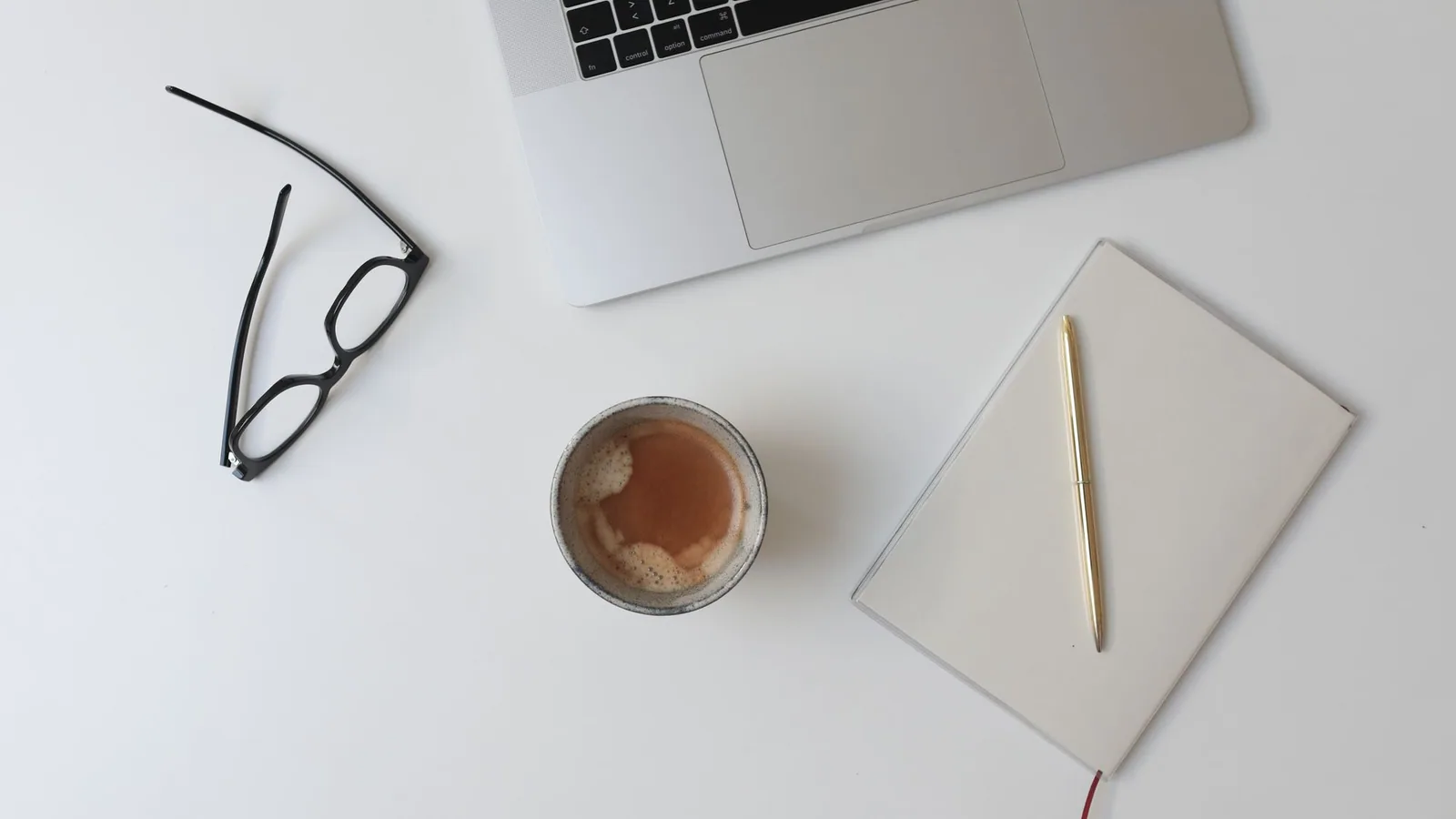 A notebook, laptop and coffee arranged on a white desk.